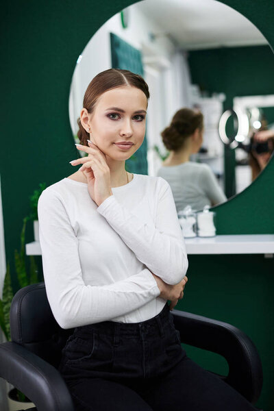 Young girl with fancy hairdo and make-up posing in front of mirror in beauty studio. Portrait of makeup fashion model. Love yourself female concept. Bride's morning on wedding day.