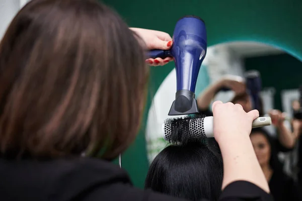 若いブルネットの女性はパーティーの準備をしている 大きな鏡の前にヘアドライヤーと女性のクライアントのためのヘアスタイルを作るプロの美容師 理容室での作業工程のクローズアップ写真 — ストック写真