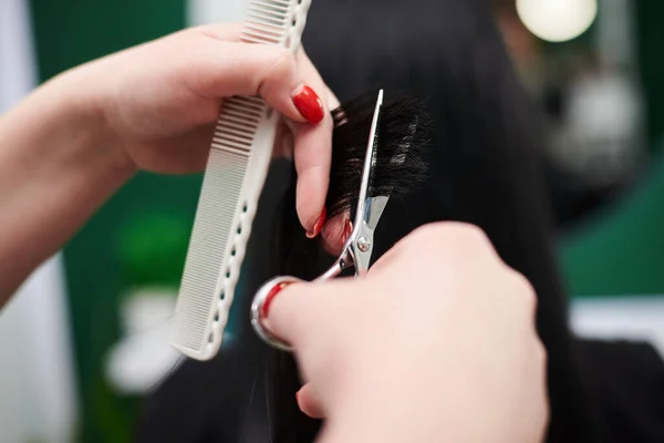 女性のクライアントのためのヘアカットを作るプロの美容師 ヘアスタイリストの手にはさみや櫛を持ち 光沢のある黒い髪を切断するのクローズアップ画像 理容室での作業工程 — ストック写真