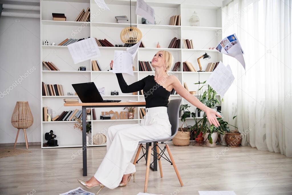 Young blond woman, wearing black top and white pants, sitting on modern grey chair, throwing papers in the air.Office manager at her workplace. Female working in company. Busy workday.