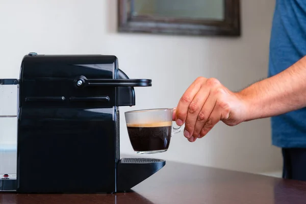 Close up of hand picking up a cup of espresso of capsule coffee machine at home. Concept of coffee break.