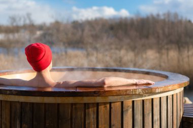 Genç yetişkin, dışarıda, tahta jakuzide dinleniyor ve doğayı seyrediyor. Güneşli bir günde sıcak havuzun keyfini çıkaran kişi, özel spa tedavisi