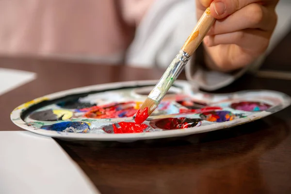 Close up of young artist hand dipping paint brush into red paint. Creativity and expression for kids.