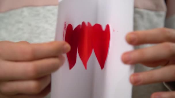 バレンタインデーに手作りグリーティングカードを展開する手 白い紙に赤いペンキでペイントしたサインが好き — ストック動画
