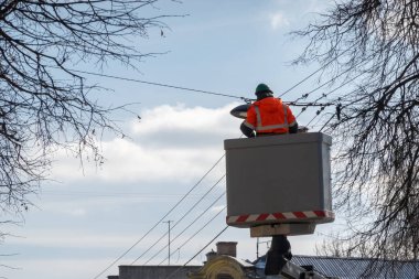 Güneşli bir günde asansör platformunda koruyucu iş kıyafetleri giyen elektrikçi sokak lambalarını değiştiriyor..