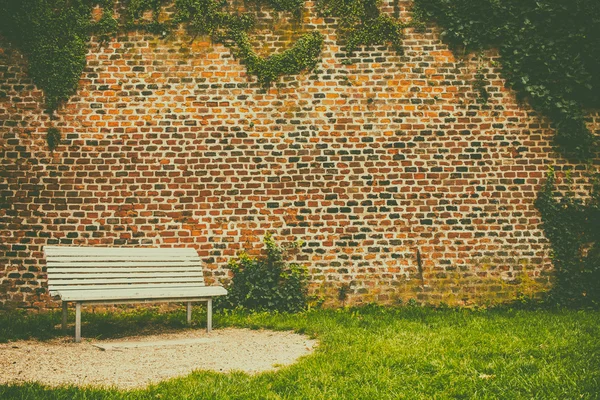 Banco blanco de pie frente a una vieja pared de ladrillo en un parque de la ciudad . — Foto de Stock