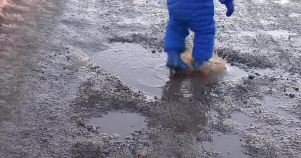 Child in bright blue overalls and rubber boots walk and jump in muddy puddle of black water and earth. Concept about childhood and dirty clothes, holes in roads. — Wideo stockowe