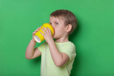 Aç gözlü çocuk arkasına dönüp yeşil arka planda sarı tek kullanımlık fincandan kahve içiyor. Çocukluk kıskançlığı ve insan psikolojisi kavramı.