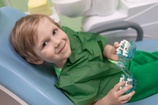 Улыбающийся кавказский ребенок исследует зубы в челюсти на стуле у стоматолога крупным планом. Концепция детской стоматологии, игр и игрушек в клинике.