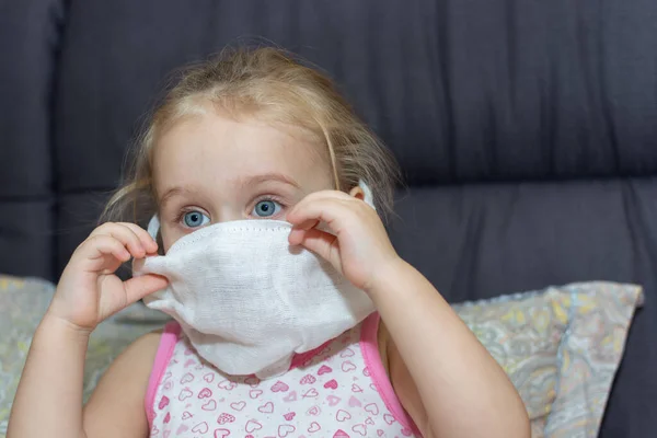 Little girl takes off the medical gauze mask from her face while sitting on the bed at home. End of home treatment.