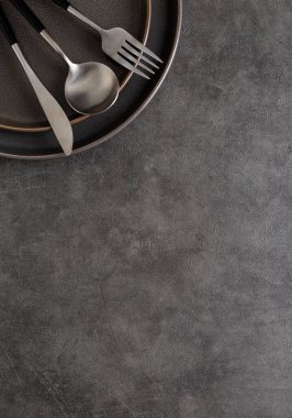 Çatal, kaşık ve bıçak koyu yuvarlak seramik plaka üzerinde koyu gri beton arka plan ve fotokopi alanı. Üst aşağı görünüm, düz görünüm, metin için yer, şablon