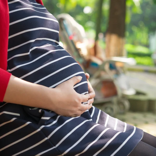 年轻孕妇把双手放在腹部感觉婴儿，婴儿小推车上方形组成的背景下， — 图库照片