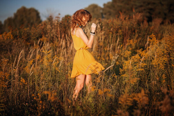 Stylish girl in a yellow dress dancing among the grasses and wildflowers in a sunny meadow in the mountains.