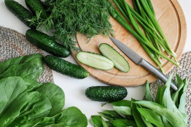 Sliced fresh cucumbers in a bowl, green onions, dill, and on a cutting board. Ingredients for a summer salad on a white tree.