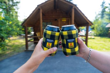 Montana, USA - July 2, 2021: Two campers at a KOA (Kampgrounds of America) use koozies or can cozy to toast to a fun summer vacation clipart
