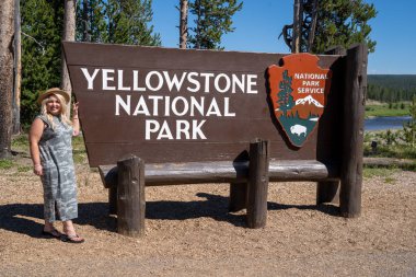 Wyoming, ABD - 28 Haziran 2021: Yellowstone Natinoal Park giriş tabelasının yanında fötr şapka takan mutlu sarışın kadın