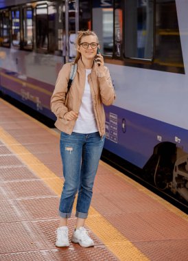 Trenin yanındaki platformda gözlüklü kadın. Mavi trenin yanındaki tren istasyonunda cep telefonu olan bir kız. Demiryolu ile seyahat et. Bydgoszcz.