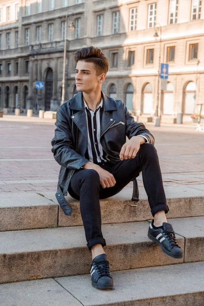 Stylish guy sitting on the street. Young man model appearance. A man in a black smoker and satan and a black and white shirt. Photo shoot on the street in Warsaw. Poland.