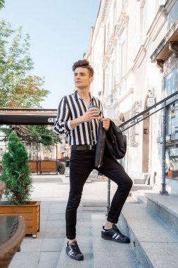 Sokakta bir bardak sıcak içeceği olan genç bir adam. Yakışıklı bir adam merdivenlerden yukarı çıkıyor. Kolunda bir fincan kahve ve siyah bir ceket olan adam. Seyahat.