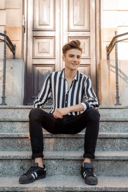 Genç adam evin önündeki merdivenlerde oturuyor. Şık giyinmiş şık bir adam. Eski şehrin sokaklarındaki genç İtalyan adam. Merdivenlerde poz veren erkek manken..