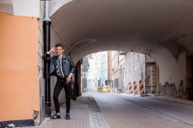 Siyah ceketli çekici genç hippi. Şehirde yürür. Yürüyüşe çıkmış çekici bir adam. Kentin yeni portresi. Kendine güvenen, saç stili, dışarıda duvarın yanında siyah kot ceket giyen genç adam..