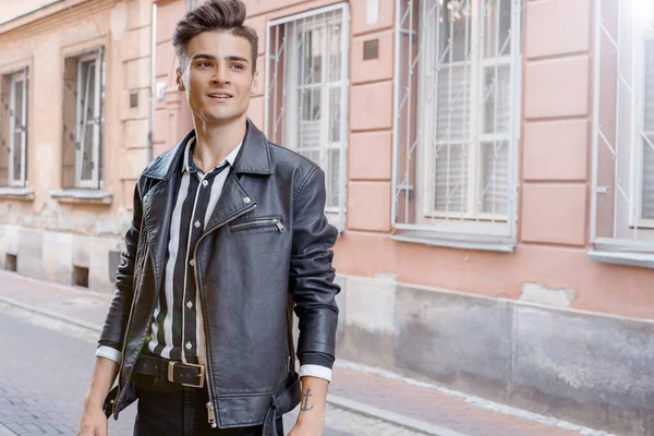 Attractive young hipster man in black jacket. walks in the city. Attractive guy on walk. Urban fresh portrait confident young man with hairstyle in fashion black denim jacket near wall outdoors.