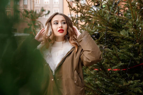 ニットのセーターとふわふわのヘッドフォンの女性。若い美しい幸せな笑顔の女の子の屋外肖像画は、装飾されたクリスマスツリーの近くに毛皮のコートを着ている. — ストック写真