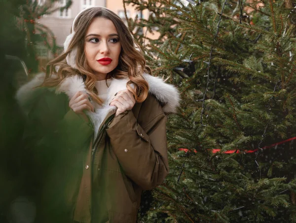 ニットのセーターとふわふわのヘッドフォンの女性。若い美しい幸せな笑顔の女の子の屋外肖像画は、装飾されたクリスマスツリーの近くに毛皮のコートを着ている. — ストック写真