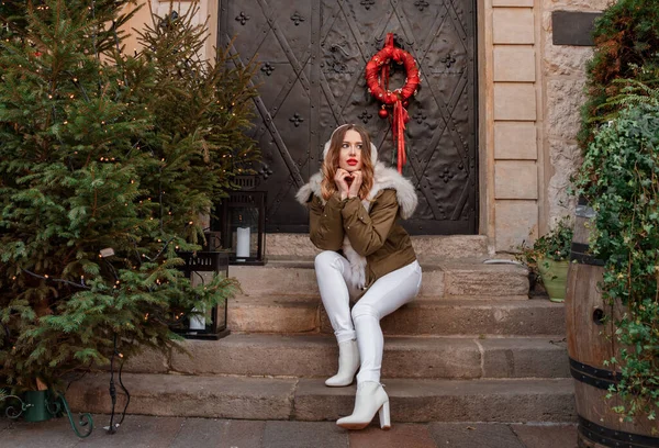 Vacker ung kvinna sitter på trappan nära julgranen. En söt flicka tillbringar tid på julmarknaden i julen. Porträtt av charmig ung kvinna tittar på kameran glatt håller. — Stockfoto