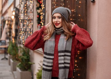 Şık örülü şapka ve eşarp takıyordu. Kadın, arka planda ışıklar, dekorasyonlar ve atlıkarıncayla Noel pazarında yürüyor..