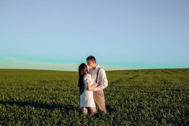 Birbirlerine aşık güzel bir çift yeşil bir tarlada çalılarla oturup sarılıyorlar. Güneşli bir yaz gününde yeşil arazide yürümek. Ülkedeki doğa. Şapkalı kız ve erkek..