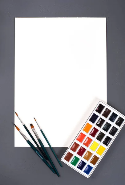 Pintura para desenho e paleta em um fundo cinza. Uma folha de papel branca em branco. — Fotografia de Stock