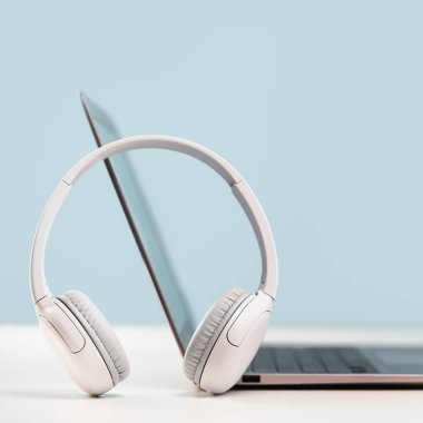 Gri masadaki kablosuz kulaklıklı ince dizüstü bilgisayarın yan görüntüsü. Mavi arka plan. Uzaktan öğrenme. Evden, çevrimiçi kurslardan ya da destekten çalışmak. Ses yayını. Yardım masası veya çağrı merkezi kulaklığı