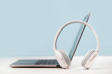 Gri masadaki kablosuz kulaklıklı ince dizüstü bilgisayarın yan görüntüsü. Mavi arka plan. Uzaktan öğrenme. Evden, çevrimiçi kurslardan ya da destekten çalışmak. Ses yayını. Yardım masası veya çağrı merkezi kulaklığı