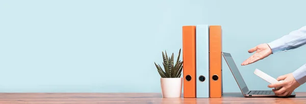 Slim laptop. Hands of female business woman pointing at screen holding headphones. Blue and orange document binders files on an neat office desk. Cactus in a pot. Business concept banner. Copy space