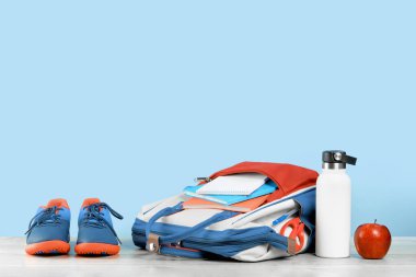 Okul kırmızı ve mavi kırtasiye malzemeleriyle dolu sırt çantası, gri ahşap masada kırmızı elmalı spor ayakkabıları. Su şişesi. Okul eğitimi ve spor konsepti maviye dönmek için alanı kopyala