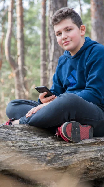 春の午後 少年は携帯電話を手に森の中の丸太の上に座る — ストック写真