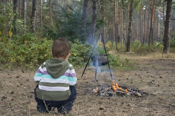 El niño se sienta en el bosque cerca del fuego, donde la comida es coo — Foto de Stock