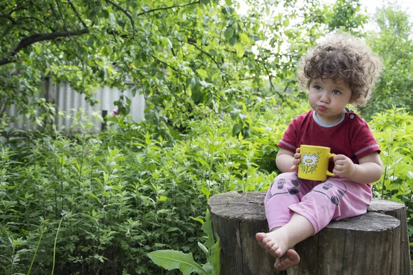 Im Sommer sitzt im Garten ein kleines, lockiges Mädchen und hält eine Tasse — Stockfoto