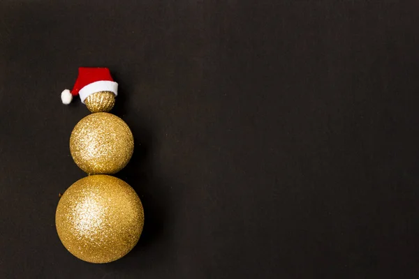 Jul snögubbe gjord av gyllene julbollar i olika storlekar på en svart bakgrund. Röd tomteluva på en snögubbe. Julkreativt. Kopiera utrymme. — Stockfoto