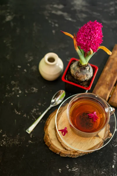 A cup of tea in a transparent cup with spring hyacinth flower on a black background. The mug stands on a wooden frame, next to it lies a metal spoon and a white vase. Copy space — Stock Photo, Image