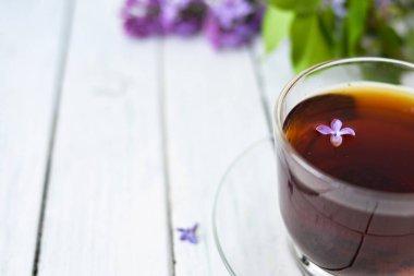 Sabah sehpasında leylak çiçekleri, şeffaf bir fincan çay ve bir fincan çay tabağı ve beyaz tahta üzerinde beyaz bir vazo. Uzayı kopyala Bahar kahvaltısı kavramı.