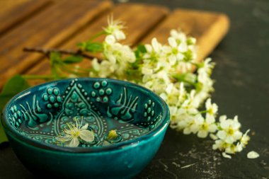 Ahşap bir tahtada beyaz kiraz çiçeklerinin arka planında şeffaf su bulunan mavi bir Türk kasesi ve siyah bir arka plan. Uzayı kopyala Bahar tatili kavramı