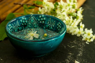 Ahşap bir tahtada beyaz kiraz çiçeklerinin arka planında şeffaf su bulunan mavi bir Türk kasesi ve siyah bir arka plan. Uzayı kopyala Bahar tatili kavramı