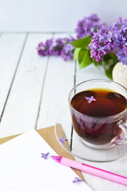 Leylak çiçekleri, şeffaf bir fincan çay ve bir fincan çay, beyaz bir vazo ve notlar için el işi kağıt ve beyaz ahşap bir masanın üzerinde pembe bir kalem. Modelleme. Uzayı kopyala Bahar kahvaltısı kavramı.