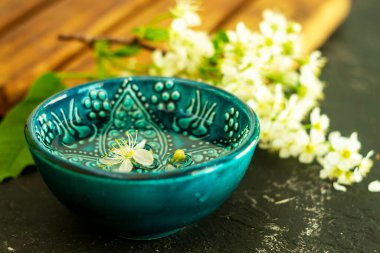 Ahşap bir tahtada beyaz kiraz çiçeklerinin arka planında şeffaf su bulunan mavi bir Türk kasesi ve siyah bir arka plan. Uzayı kopyala Bahar tatili kavramı