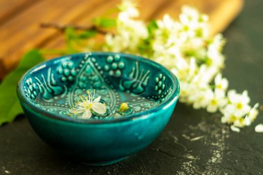 Ahşap bir tahtada beyaz kiraz çiçeklerinin arka planında şeffaf su bulunan mavi bir Türk kasesi ve siyah bir arka plan. Uzayı kopyala Bahar tatili kavramı