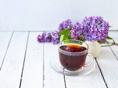 Sabah sehpasında leylak çiçekleri, şeffaf bir fincan çay ve bir fincan çay tabağı ve beyaz tahta üzerinde beyaz bir vazo. Uzayı kopyala Bahar kahvaltısı kavramı.
