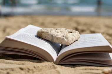 Sahilde kumsalda bir taş tarafından ezilmiş açık bir kitap yatıyor. Arka plan bulanık. Plajda yaz tatili.