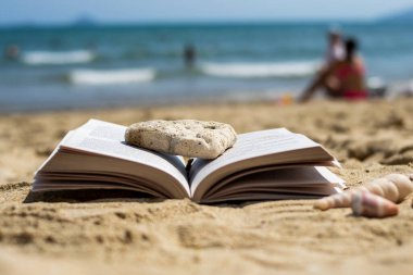 Sahilde kumsalda bir taş tarafından ezilmiş açık bir kitap yatıyor. Arka plan bulanık. Plajda yaz tatili.